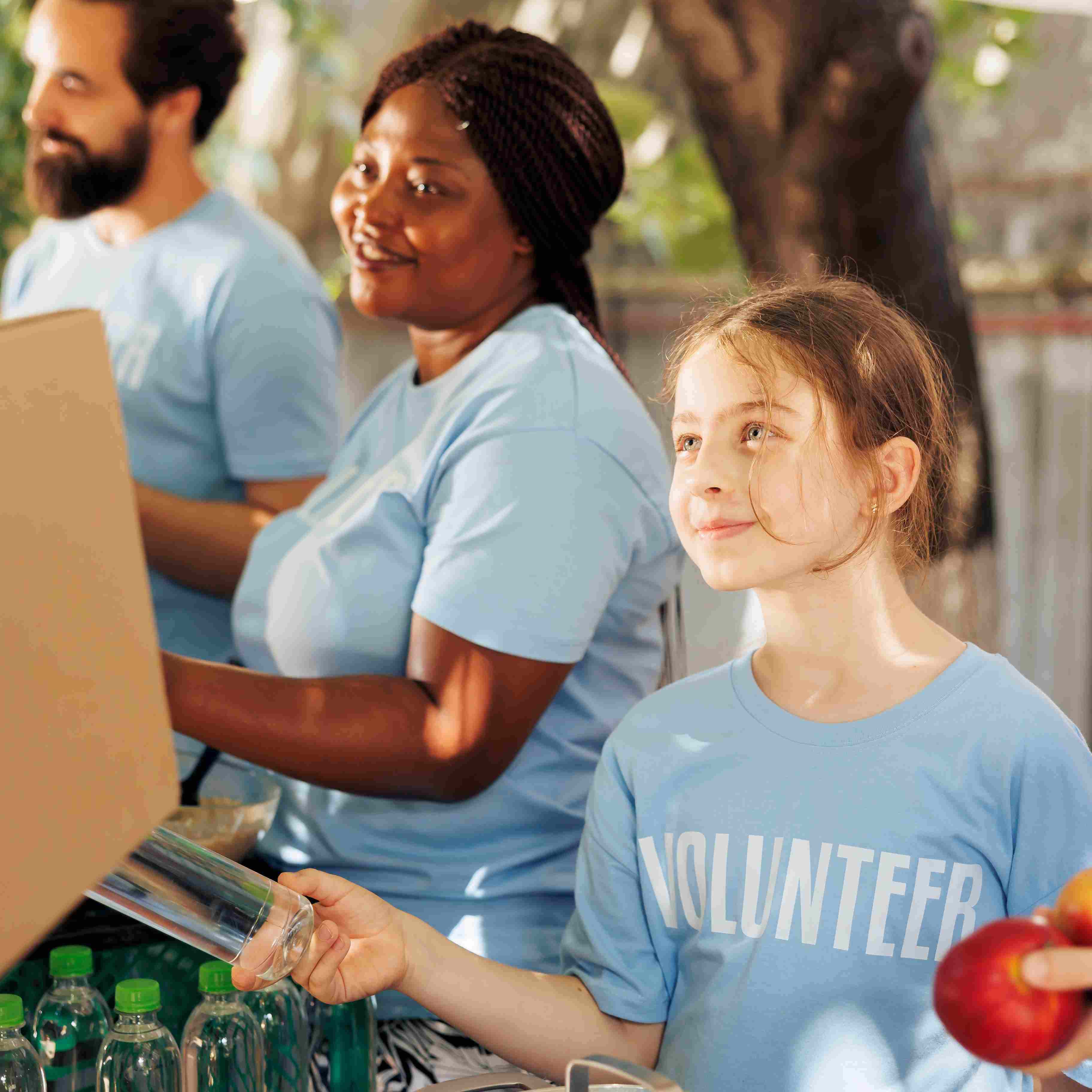 Three people wearing blue shirts, two of whom are holding bottles and an apple, with a blurred outdoor background.