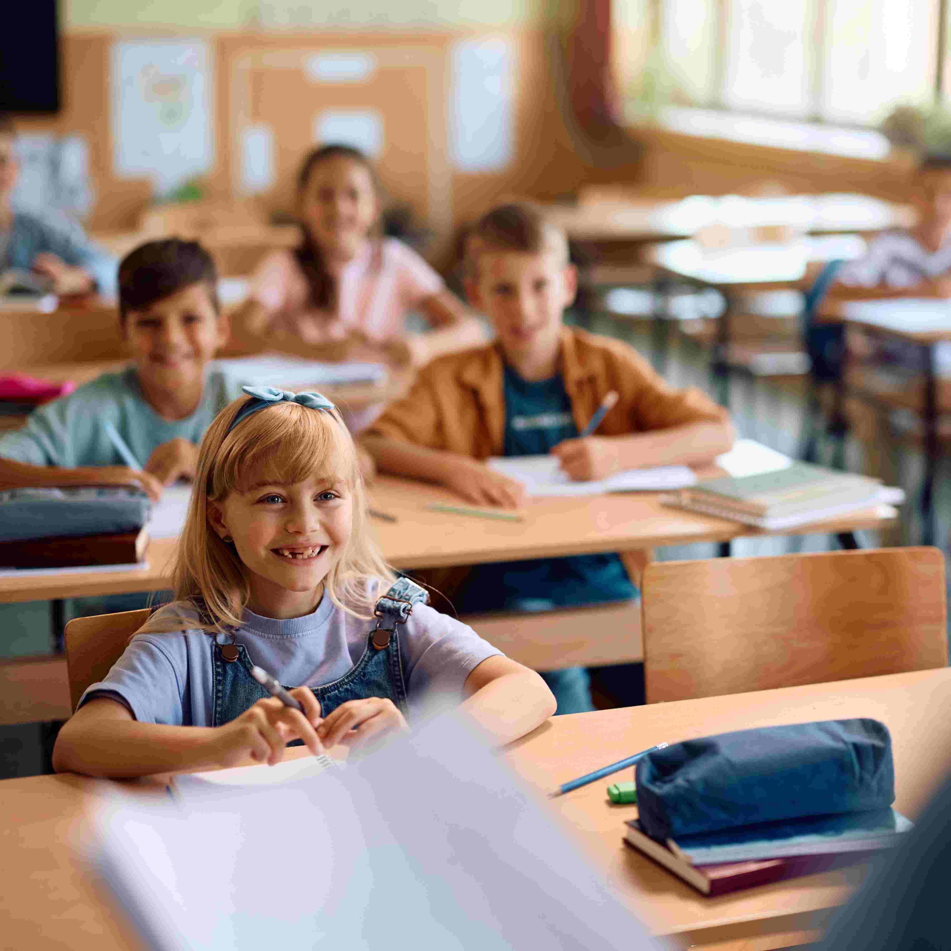 Children sitting at desks in a classroom, with one girl smiling at the camera.