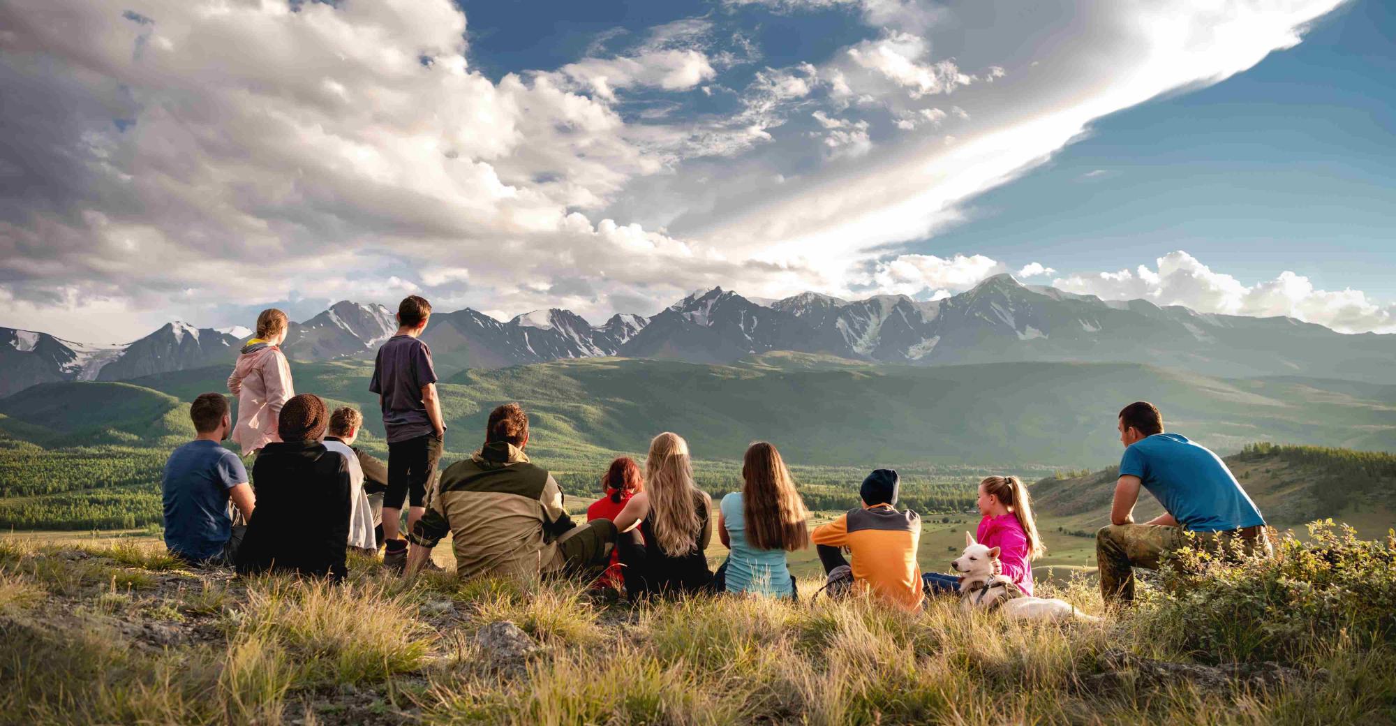 Group of people sitting on a grassy hill overlooking mountains with a rainbow in the sky.