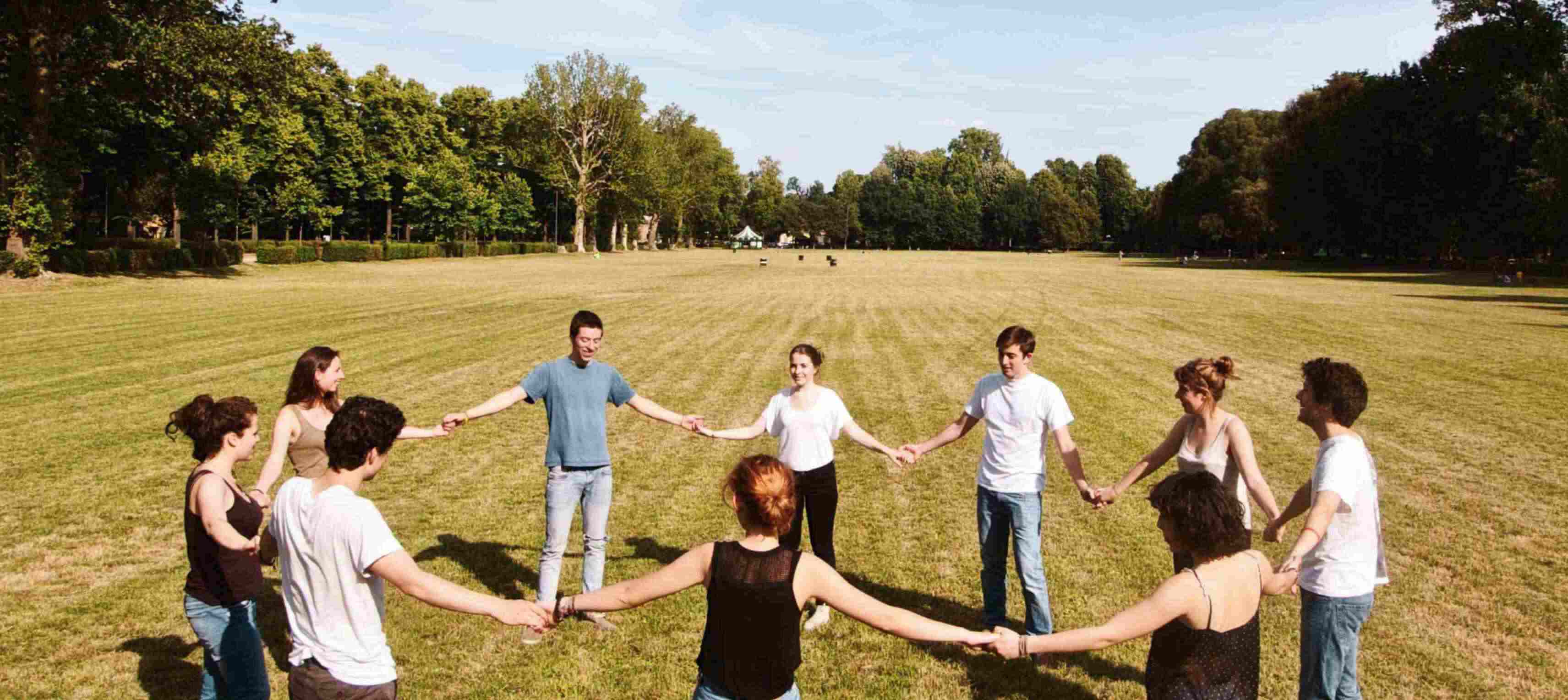 Group of people holding hands in a circle on a grassy field with trees in the background